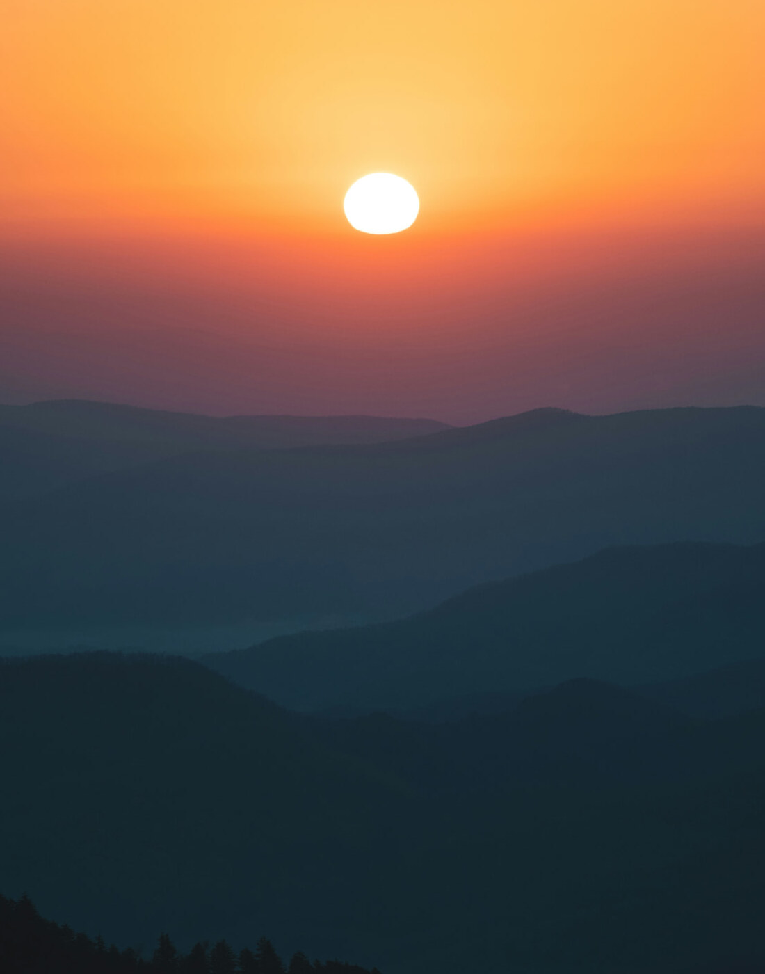 Sun setting over layered blue mountain ridges beneath a golden orange sky